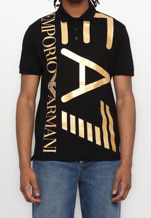 Black polo shirt featuring large gold text and graphic accents. Made from cotton, with a collar and short sleeves.