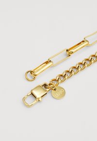 Accessoire en chaîne dorée avec des maillons entrelacés, un fermoir et une étiquette ronde gravée "LJUJO". Texture métallique lisse et brillante.