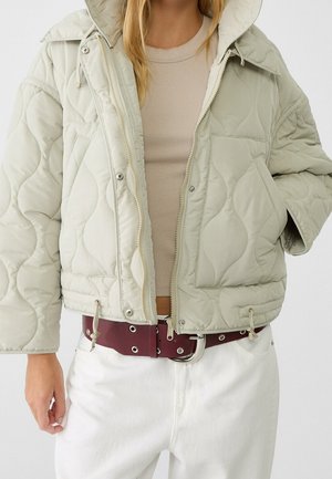 Kvinde iført en lysebeige quiltet hættetrøje, beige ribbet top, hvide bukser og et bredt mørkerødt bælte med sølvøjer og spænde.