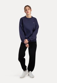 Felpa blu navy a girocollo con vestibilità leggermente oversize, abbinata a pantaloni jogger neri e scarpe da ginnastica bianche. Design semplice con branding minimale.