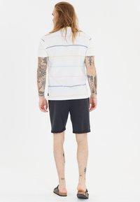 Lyhythihainen valkoinen polo-paita, jossa on monivärisiä vaakasuoria raitoja, yhdistettynä tummansinisiin shortsihin. Kädet ovat tatuoituja ja jaloissa on kevyet kengät.