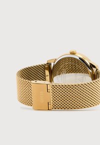 Gouden gaas horloge met een ronde kast en zilveren achterkant. Beschikt over een tekstuuroband, een sluiting en een gegraveerd merklogo op de sluiting.