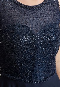 Laivansinis mekko, jossa on paljeteilla koristeltu bodice, jossa on teksturoitu, kiiltävä pinta, V-mallinen kaula-aukko ja sileä hame.