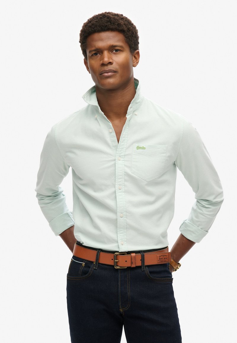 Licht muntgroen overhemd met knopen en een borstzakje en logo. Gecombineerd met donkere jeans en een bruine leren riem met een gouden gesp.