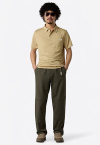 Polo beige con logo en el pecho y mangas cortas, combinado con pantalones verde oscuro y zapatos marrón claro, complementado con gafas de sol oscuras.