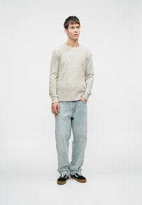 Suéter de manga longa canelado cinza claro combinado com jeans azuis claros de corte solto e ténis pretos com detalhes brancos. Fundo simples.