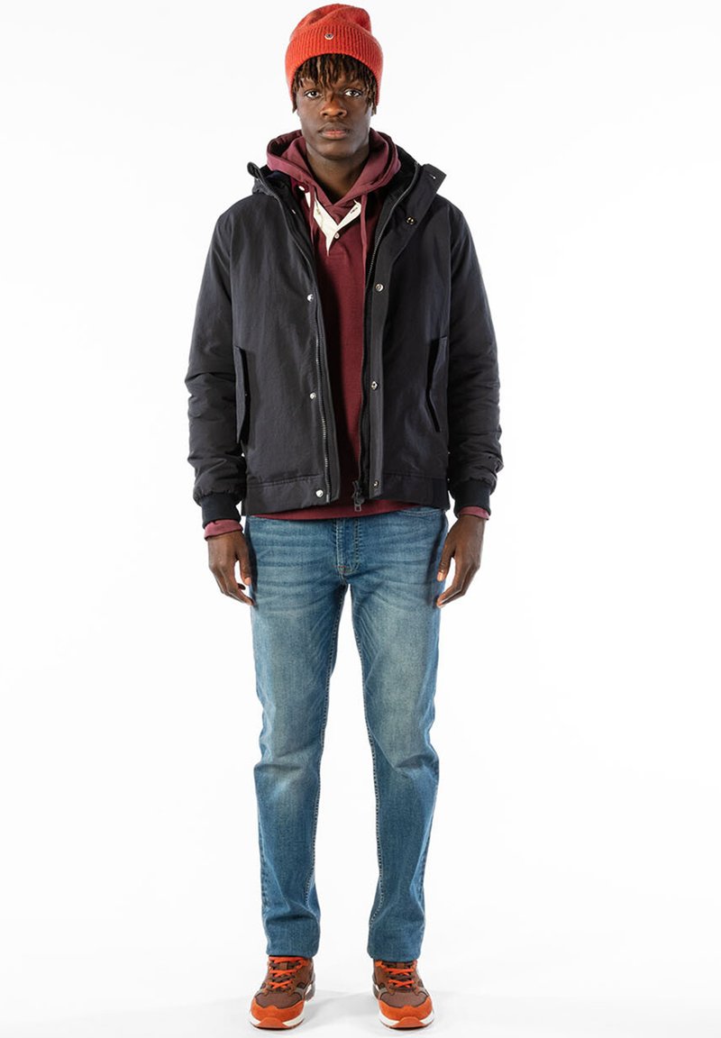 Veste à capuche noire avec fermeture éclair, sweat à capuche bordeaux en dessous, jeans bleus et sneakers avec accents orange. Le modèle porte un bonnet en tricot rouge.