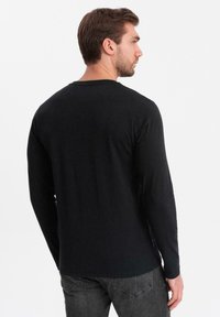 Hombre con pelo castaño corto, vistiendo una camiseta de manga larga negra lisa y jeans gris oscuro, de espaldas frente a un fondo blanco.