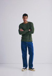 Zelený žebrovaný pletený svetr, modré džíny rovného střihu a černé tenisky s bílou podrážkou. Model stojí s překříženýma rukama na světlém pozadí.