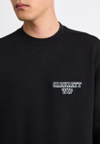 Black sweatshirt made of cotton, featuring ribbed neckline and cuff. "Carhartt WIP" logo embroidered in white on the left chest.
