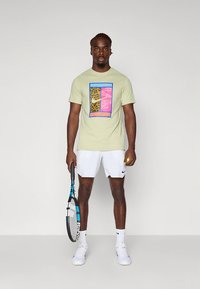 Homem em pé segurando uma raquete de ténis e uma bola, vestindo uma t-shirt gráfica bege, calções brancos, meias brancas e sapatilhas de ténis brancas.