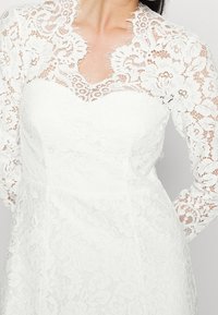 Valkoinen pitsimekko pitkillä hihoilla, jossa on aaltoileva kaulus ja kukkakuosisia yksityiskohtia, päällystetty tiukasti istuvalla yläosalla, tekstuurinen kangas.