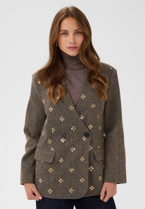 Femme aux longs cheveux bruns portant un col roulé marron et un blazer marron croisé à petits motifs fleuris dorés, debout sur un fond gris.