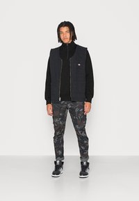 Gilet di jeans nera sopra un top nero con zip, abbinato a pantaloni cargo mimetici scuri e sneaker alte nere con strisce bianche.