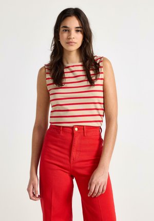 Ung kvinde med langt mørkt hår iført en ærmeløs beige top med røde striber og højtaljede røde bukser mod en ensfarvet baggrund.