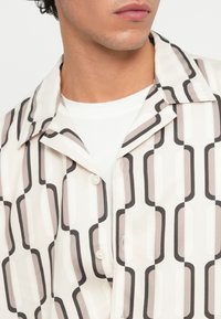 Camicia a maniche corte con un motivo geometrico in tonalità beige, grigio e nero. Presenta un colletto a bottonatura e una chiusura con bottoni. Tessuto dalla texture liscia.