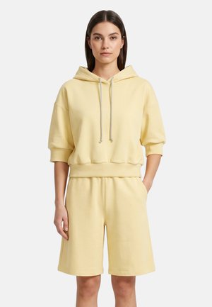 Vrouw draagt een lichtgele capuchonsweater met driekwart mouwen en bijpassende knielange shorts, staat met één hand in de zak.