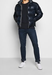Chaqueta acolchada en azul marino, con una superficie texturizada y cierre con cremallera. Combinada con jeans oscuros y zapatillas blancas.