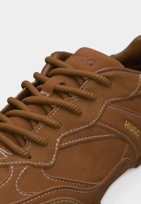 Καφέ σουέντ αθλητικό παπούτσι με χονδρές κορδόνια, λευκή ραφή και λογότυπο "HUGO" σε χρυσό στην πλευρά.