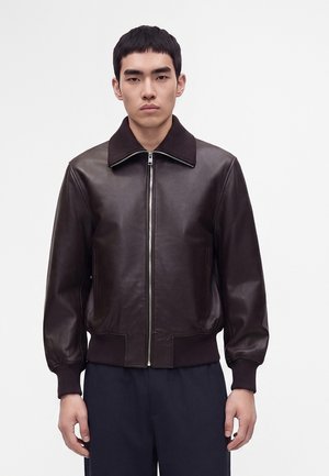 Jeune homme debout, face à l'avant, portant une veste zippée en cuir marron foncé avec poignets et ourlet côtelés sur un pantalon sombre.