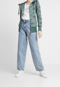 Felpa verde con motivi floreali, T-shirt bianca con grafica e jeans larghi azzurri chiari. Calzature: sneaker bianche.