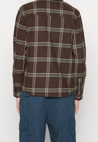 Chemise à carreaux marron avec des lignes blanches et vertes, boutonnée sur le devant, manches longues et détail de découpe au dos. Portée avec un pantalon cargo bleu.