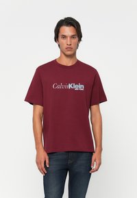 Young man wearing a burgundy Calvin Klein T-shirt and dark blue jeans, standing against a plain white background.