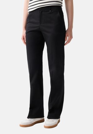 Person trägt schwarze gerade geschnittene Hose, weiße Sneakers und ein gestreiftes Hemd und steht vor einem schlichten Hintergrund.