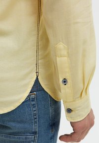 Gelbes Langarmhemd aus strukturiertem Stoff mit kontrastierenden schwarzen Nähten und knöpfbaren Manschetten, kombiniert mit blauen Jeans.