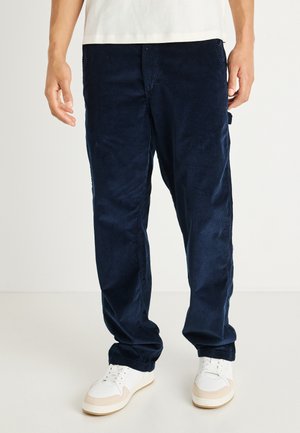 Marineblauwe corduroy broek met wijde pijpen, voorzien van verticale ribbelstructuur, zijzakken en een ontspannen pasvorm, gecombineerd met witte sneakers.