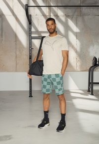 T-shirt bege com um logo preto da Puma, calções verdes com padrão, mochila de ginásio preta e sapatilhas pretas com detalhes verdes. Ambiente de ginásio.