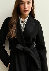 Cappotto avvolgente in misto lana nero con cintura, rever affilati e maniche lunghe. Il modello indossa una camicia a righe sotto.