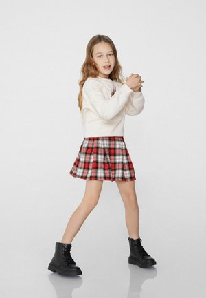 Jupe à carreaux rouge, noir et blanc ; design plissé. Associée à un pull crème clair. Porte des bottines noires à lacets.