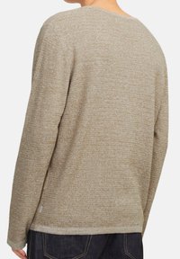 Persoon in een beige gestructureerde lange mouwen trui en donkere spijkerbroek, gezien van achteren en iets gedraaid.