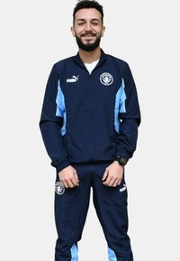 Chándal marino hecho de material ligero, con detalles en azul claro, chaqueta con cremallera y un logo, y pantalones a juego con cintura elástica.