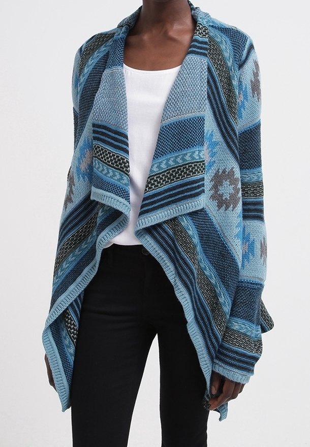 Personne portant un cardigan en maille bleu à motifs avec un col large, sur un haut blanc et un pantalon noir, visible des épaules aux hanches.
