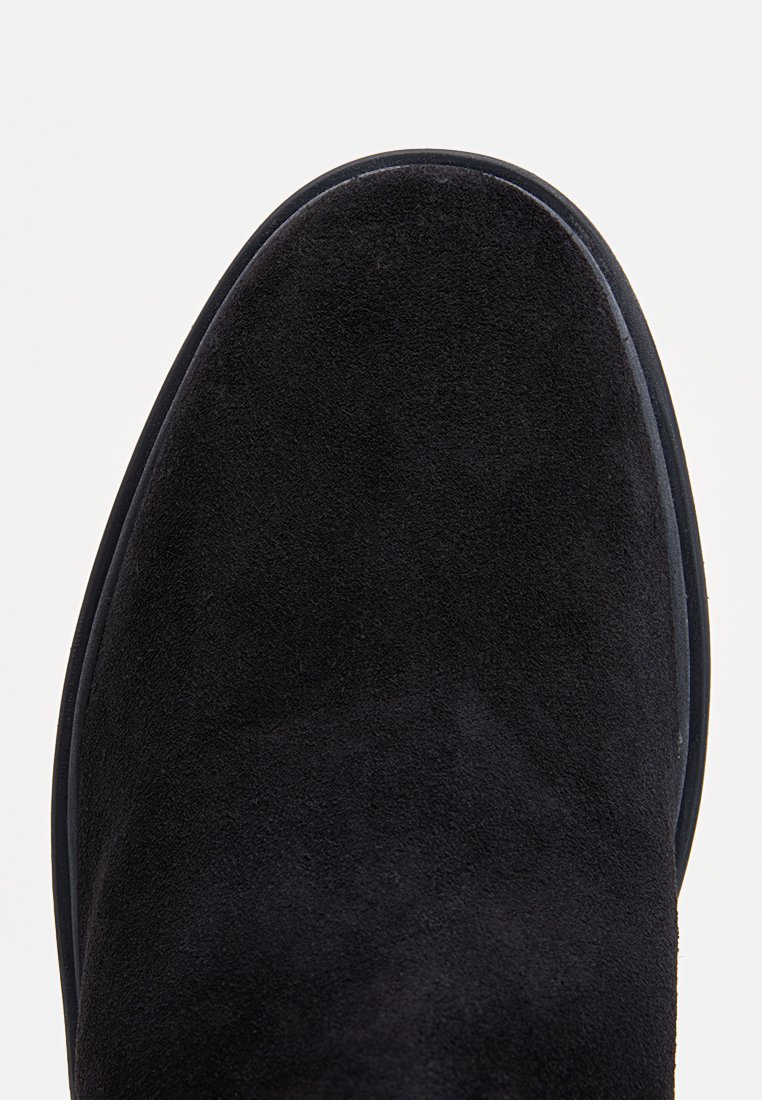 Chaussure en suede noire avec une texture lisse et un bout arrondi, présentant un léger bord en caoutchouc pour l'adhérence et la durabilité.