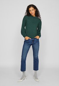 Donkergroene gebreide trui met een hoge kraag en ballonmouwen, gecombineerd met blauwe cropped jeans en lichtgrijze sokken, die puntige witte schoenen tonen.