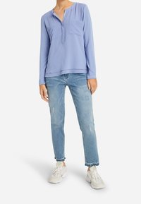 Mujer vestida con una blusa de manga larga azul claro, jeans rectos descoloridos y zapatillas blancas, de pie contra un fondo liso.