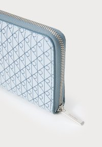 Lommebok med glidelås i lys blå med geometrisk hjerte-mønster. Har en teksturert kant og sølvfarget detalj for enkel åpning.