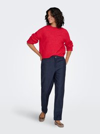 Pull tricoté rouge avec col rond, associé à un pantalon en denim foncé ample et des ballerines marron, offrant une coupe décontractée et confortable.