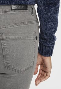 Vaqueros de mezclilla gris con un acabado texturizado, que cuentan con un bolsillo trasero con un acento triangular negro y una etiqueta de logo negra en la cintura.