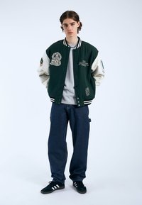 Giacca varsity verde e crema con toppe ricamate, colletto a righe e chiusura con bottoni, abbinata a jeans blu navy larghi e sneakers nere.