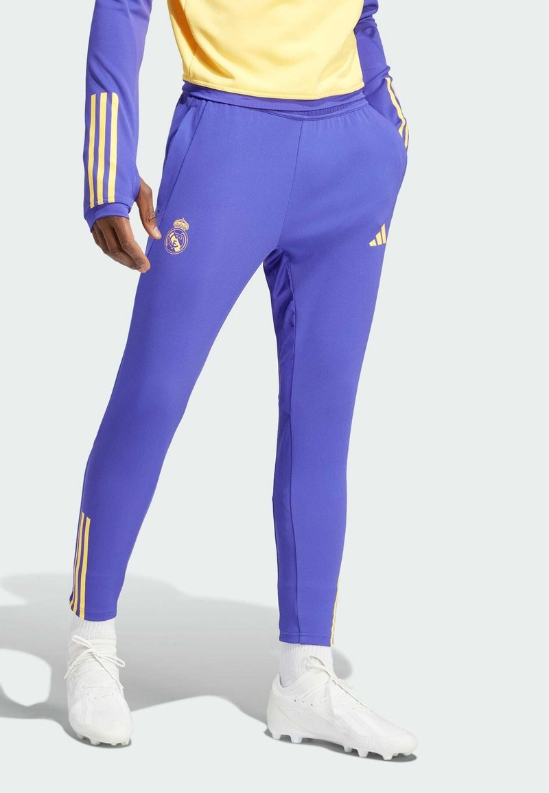 Purple athletic pants with yellow stripes, featuring a fitted design, soft texture, and a logo on the thigh, paired with white sports shoes.