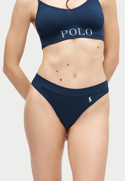 Γυναίκα που φοράει αθλητικό σουτιέν Polo σε σκούρο μπλε χρώμα και ταιριαστό σλιπ με υψηλή κοπή, με το λογότυπο Polo, σε απλό ανοιχτό φόντο.