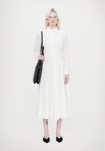 Vestido blanco con cuello y cierre de botones, que presenta un busto ajustado y una falda acampanada. Bolso negro y zapatos negros de punta.