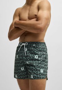 Swim shorts in green with repeated “HUGO” text pattern in black and white. Features a drawstring waistband and a lightweight material.