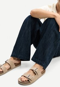 Beige mockasandaler med två justerbara spännen, korkfotbädd och texturerad gummisula, bärs med mörkblåa vida jeans.