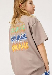 Vaaleanruskea ylipitkä t-paita, jossa on monivärinen liekkimuotoilu ja sana "COURAGE" toistuu kolme kertaa takana.