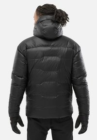 Homme avec des dreadlocks portant une veste doudoune noire isolante avec capuche et des gants noirs, tourné le dos contre un fond uni.